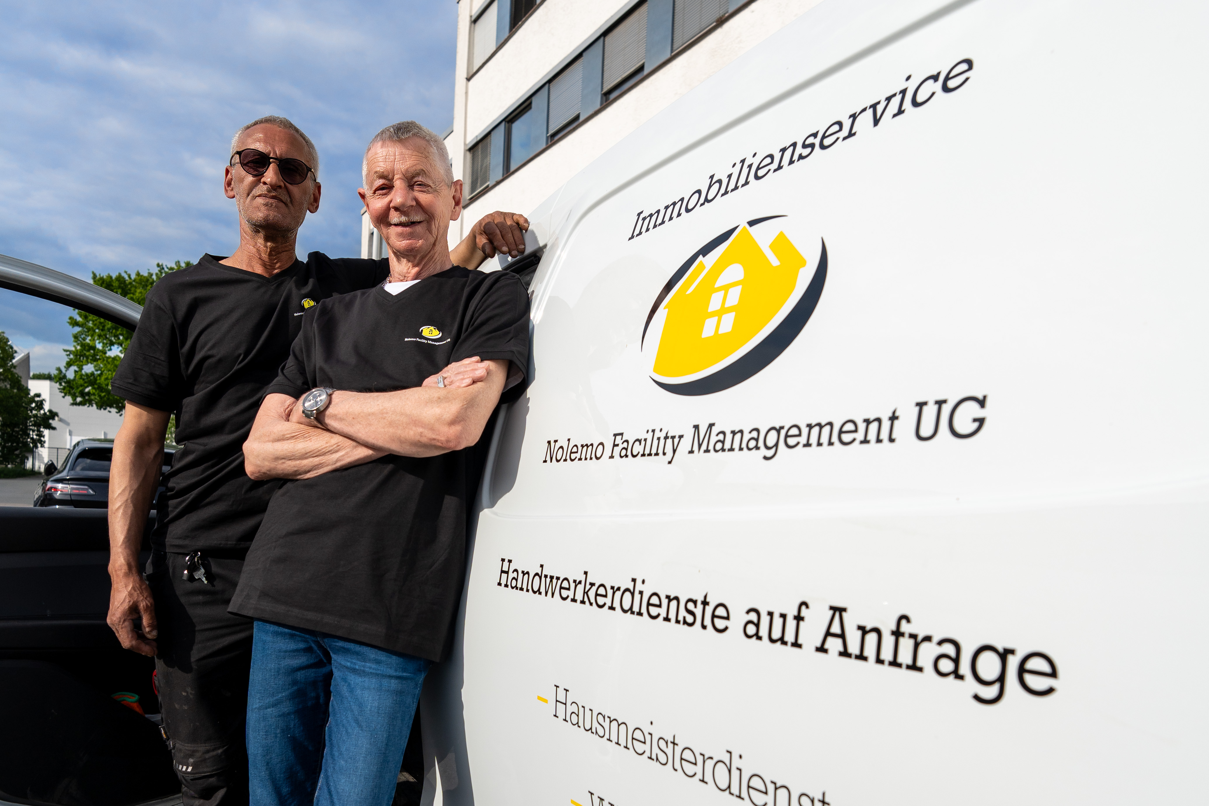 Zwei Maenner mit neutralem Blick in Schwarz neben Servicevan mit gelbem Logo